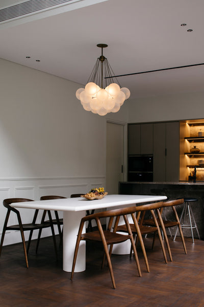 A modern white dining table with walnut dining chairs. A bubble light-fixture hangs above it and a contemporary kitchen is seen in the background. 