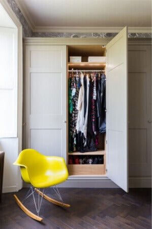 A bedroom with a herringbone wood floor with a matte finish and a yellow chair next to an open wardrobe cabinet.