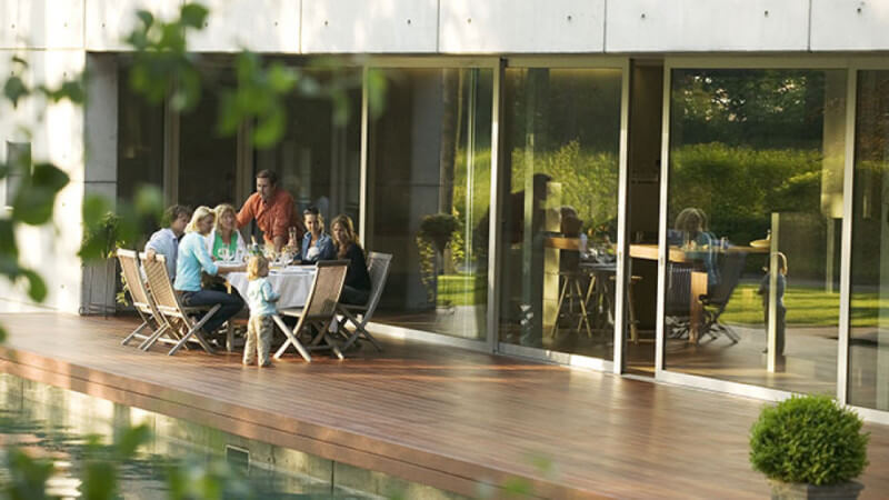 A family sitting at a table on a wood deck next to a pool and house.