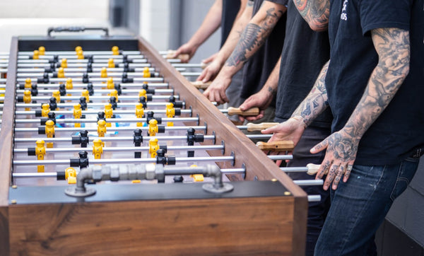 Three people are playing foosball on a walnut foosball table.
