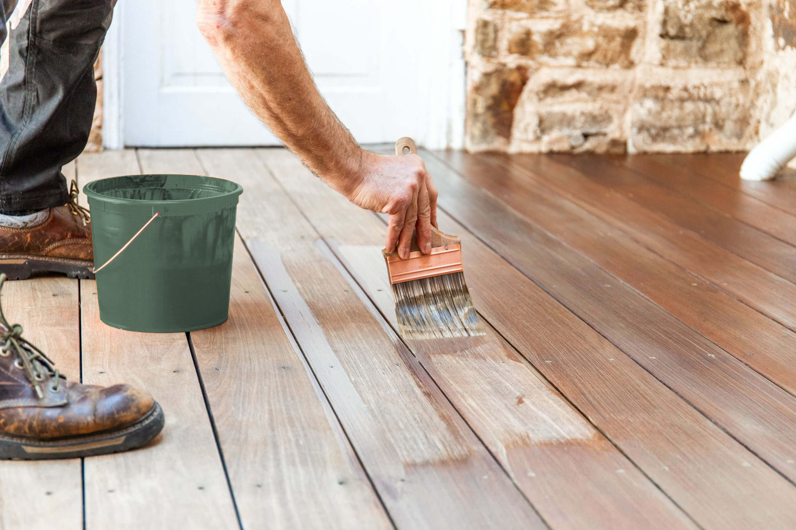 Person applying wood stain to an outdoor deck with a brush next to a green bucket
