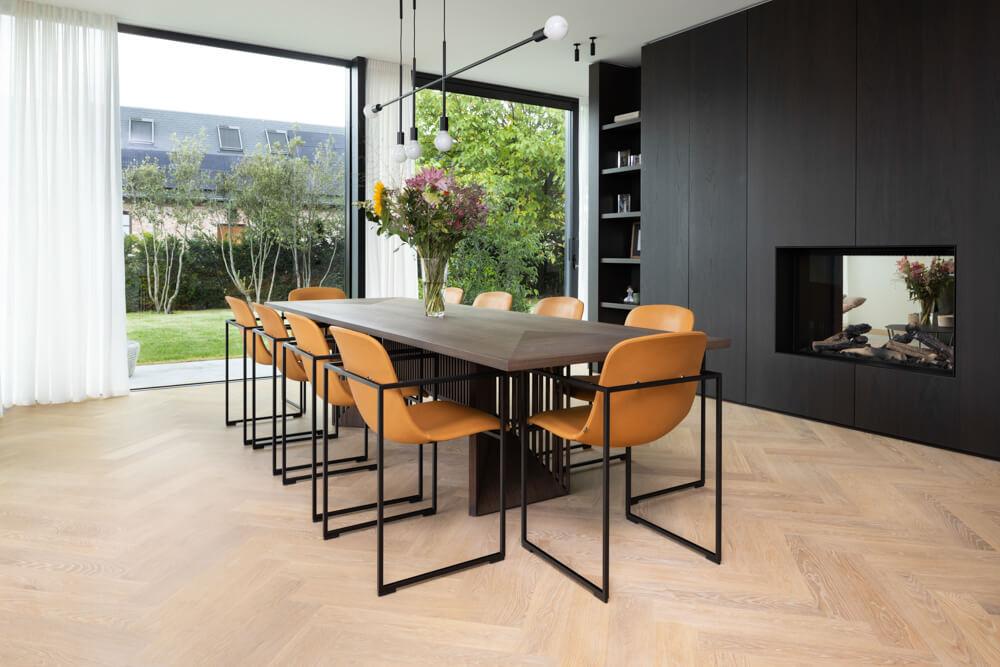 Custom herringbone flooring finished with a hardwax oil wood finish is in a dining area featuring a modern dining table with seating for ten and a dark double sided fireplace with built-in shelving.