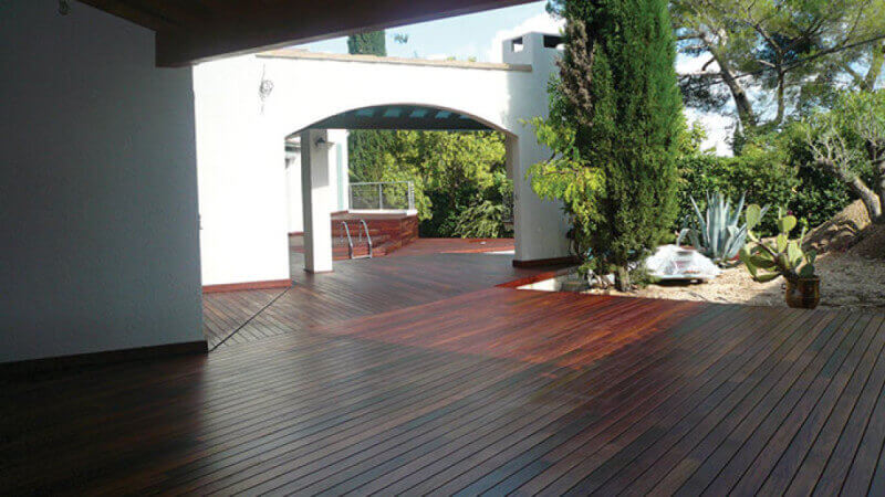 A large Ipe wood deck finished with a hardwax oil wood finish behind a house with trees and plants alongside it.
