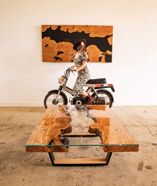 A live edge burled maple coffee table and a woman is seen riding a motorcycle in the background.