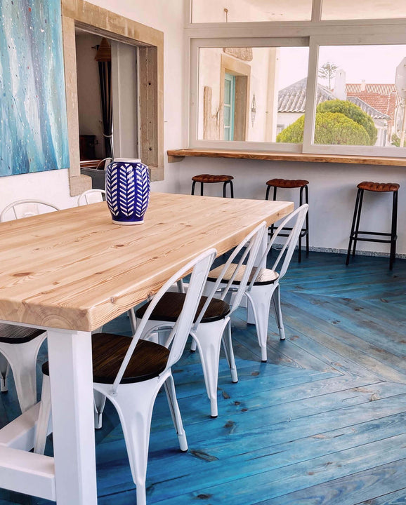 A six person wooden dining table sits in the dining room of a surf lodge. The dining room has a blue chevron wood floor.