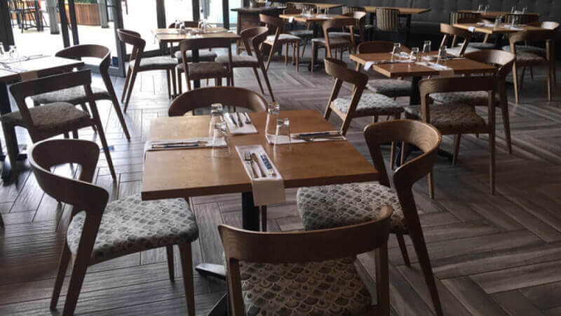 A restaurant with a herringbone style wood floor and wood tables throughout.