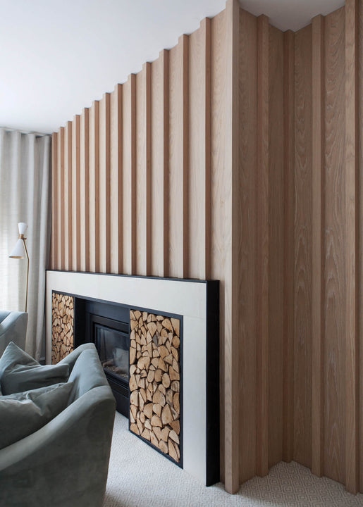 White oak slat wall finished with Rubio Monocoat Oil Plus 2C Natural wraps a room corner above a fireplace with stacked firewood storage.