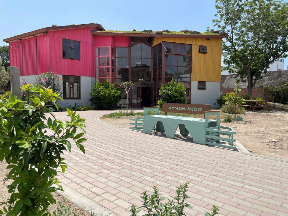 Aynimundo community building exterior with pink and yellow corrugated wood siding finished in Rubio Monocoat, a living green roof, and a teal wooden bench reading "Aynimundo" beside a brick paver path.