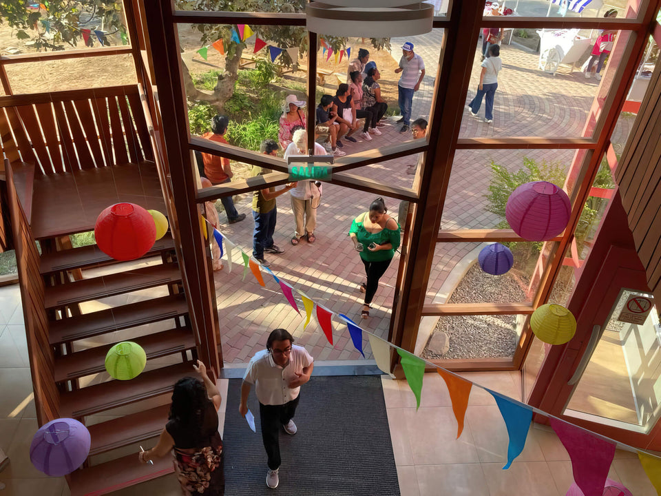 Aerial view down an open-air stairwell at Aynimundo during an event, with paper lanterns and pennant flags strung between wood beams as visitors gather on the brick plaza below.