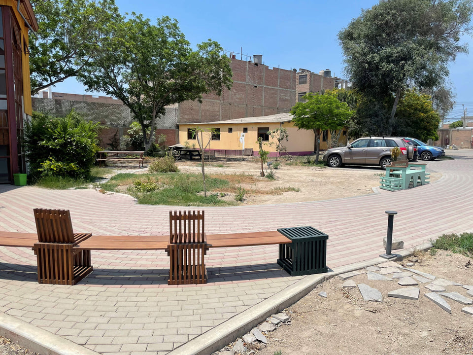 Courtyard view of Aynimundo with wood slat benches and chairs finished in Rubio Monocoat hardwax oil, a curved brick paver plaza, and the yellow-toned secondary building in the background.