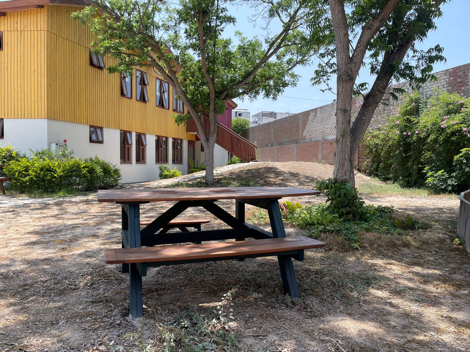 Wood picnic table with a dark stained finish sits in dappled shade near the yellow wood-clad Aynimundo building, framed by mature trees.