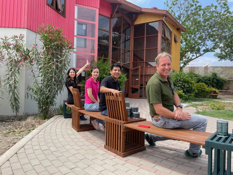 Four people sitting along a curved wood bench finished in Rubio Monocoat, with cans of Oil Plus 2C hardwax oil resting on the seat, in front of the pink and yellow Aynimundo facade.