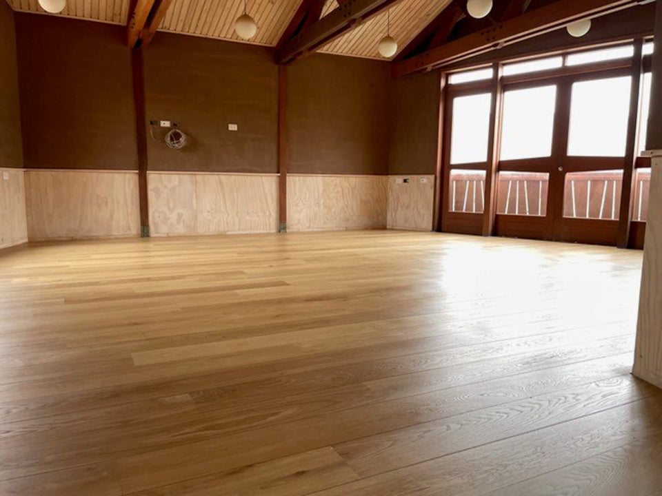 Empty interior room at Aynimundo with wide-plank oak flooring finished in Rubio Monocoat Oil Plus 2C, plywood wainscoting, exposed timber trusses, and glass doors opening to a wood balcony.