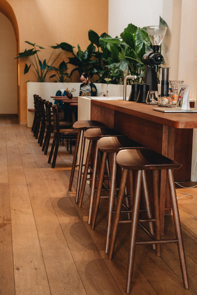 An oak counter height table has four stools resting beneath it. Several café style tables are seen in the background of the image.