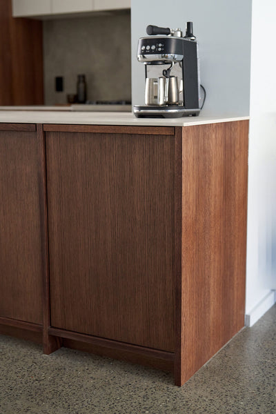 An espresso machine sits on a countertop in a modern kitchen with white oak cabinets and white countertops. 