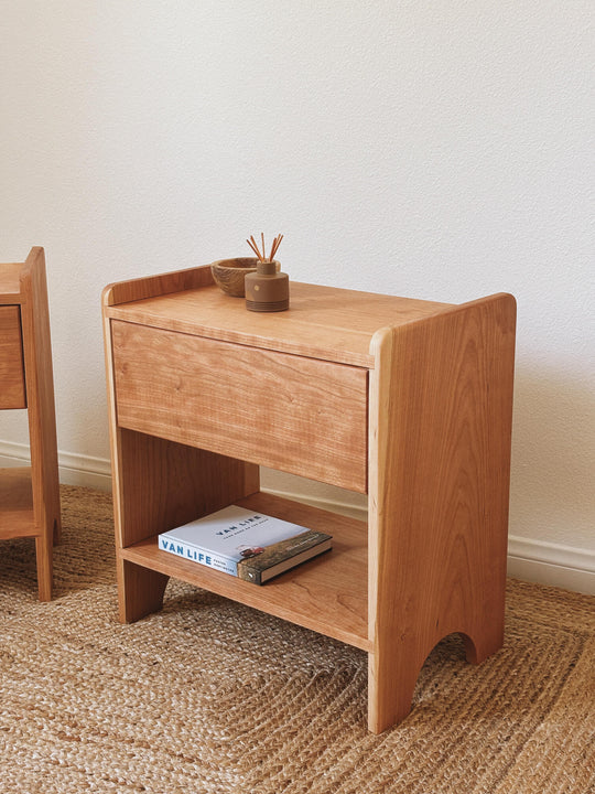Solid cherry wood bedside table with single drawer and open lower shelf, Rubio Monocoat hardwax oil Pure finish, front-facing view on woven rug.