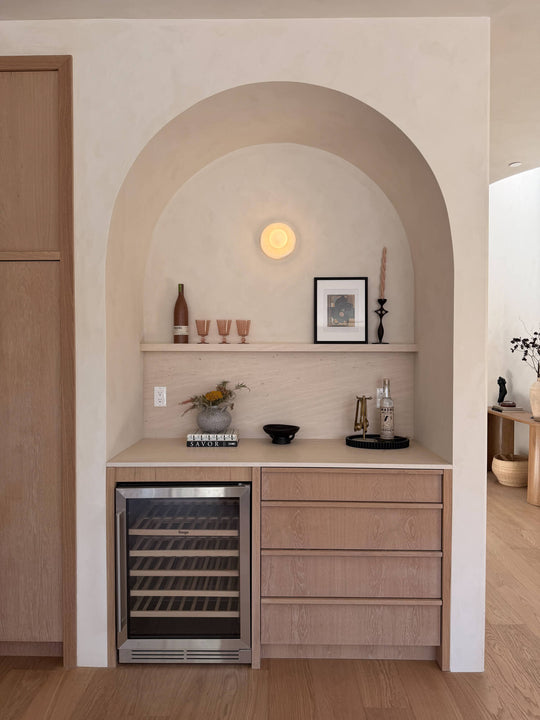 Arched wet bar niche with white oak cabinetry finished in Rubio Monocoat Oil Plus 2C Natural, wine fridge, and open shelf styled with glassware and art.