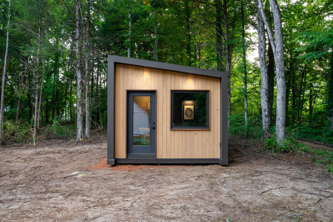 Modern backyard studio clad in vertical cypress finished with Rubio Monocoat DuroGrit in Prairie Dust, framed in dark bronze metal with a sloped shed roof, set in a wooded clearing at dusk.