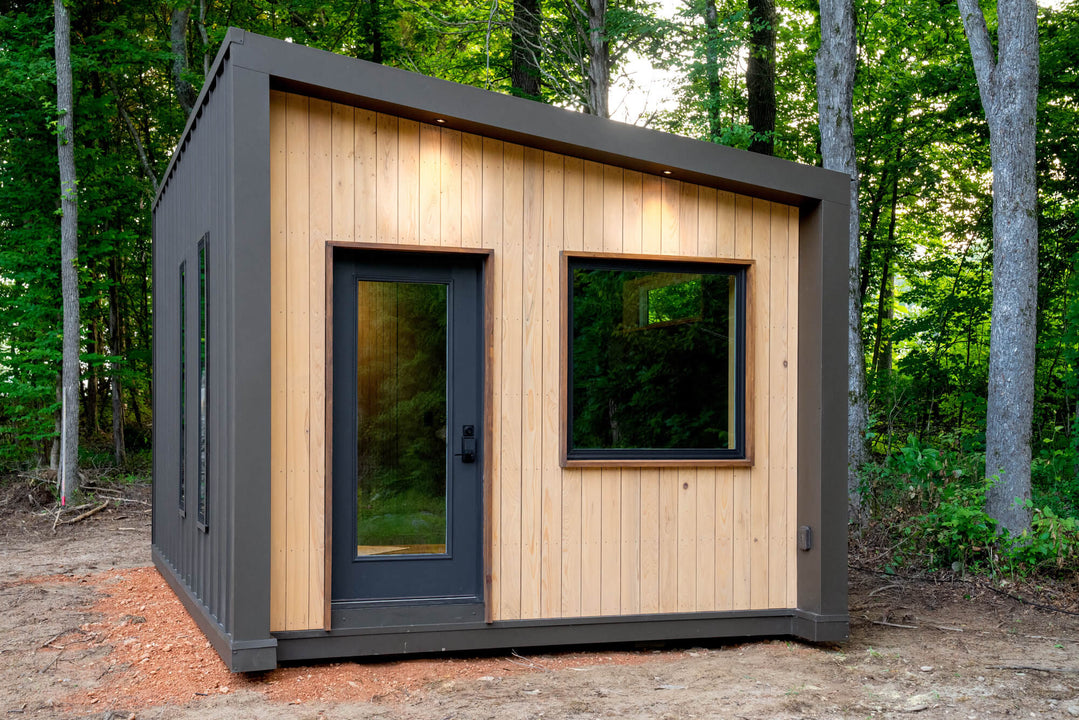 Three-quarter view of a cypress-clad studio finished with Rubio Monocoat DuroGrit Prairie Dust, with warm downlights over a black glass door and square window, and dark metal siding with narrow windows along the side.