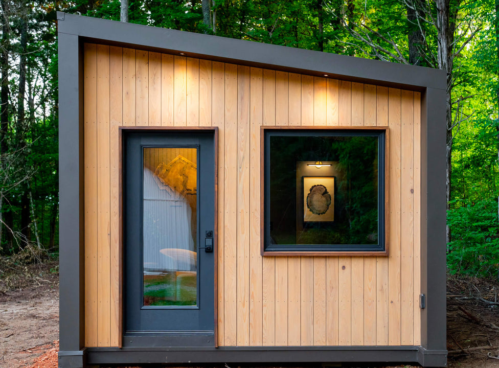 Close-up of vertical cypress cladding finished with Rubio Monocoat DuroGrit Prairie Dust, lit by recessed downlights, with a black glass door and a square window showing framed tree-ring art inside.