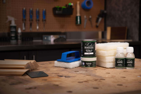 A tabletop setup of a woodworking finish kit, including a can of Rubio Monocoat oil finish, two small bottles, a blue-and-white brush, and white sanding pads, arranged on a wooden workbench with a blurred workshop background.