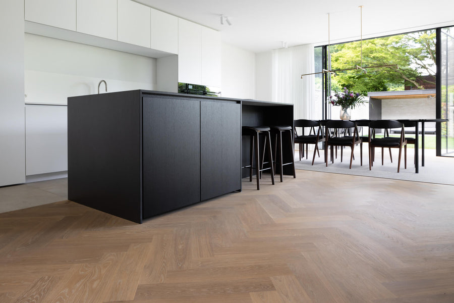 A herringbone oak hardwood floor finished with Rubio Monocoat Oil Plus 2C hardwax oil. A black wood kitchen island sits in the middle of the photos and a dining area looking to the outside is in the background.