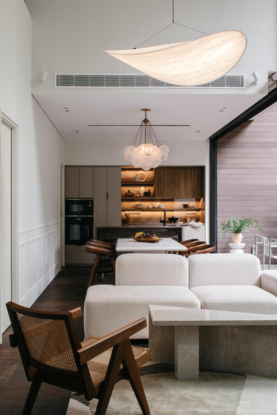 A living area with a white sofa and a brown wood chair facing the sofa. In the background you can see a modern kitchen and dining area. 