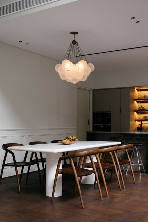 A modern white dining table with walnut dining chairs. A bubble light-fixture hangs above it and a contemporary kitchen is seen in the background.