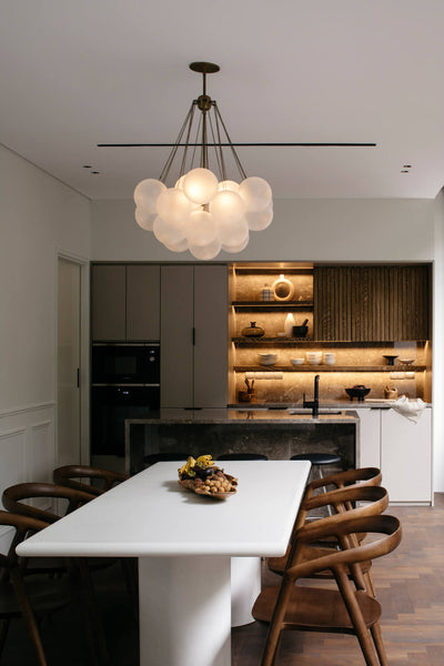 A modern rectangular dining table with six chairs is pictured here. An earth-toned, modern kitchen with dark wood cabinets and floating shelves is seen in the background. 