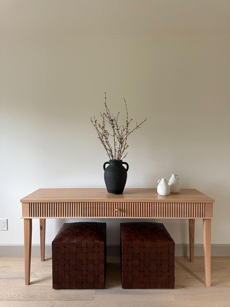 A reeded red oak console table finished in a light hardwax oil wood finish. Two stools are situated under the console table and several vases are on top.