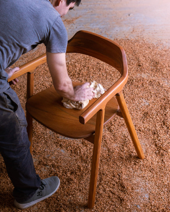 A craftsman hand-rubbing Rubio Monocoat oil finish into the seat of a walnut chair, surrounded by fresh wood shavings on a workshop floor.