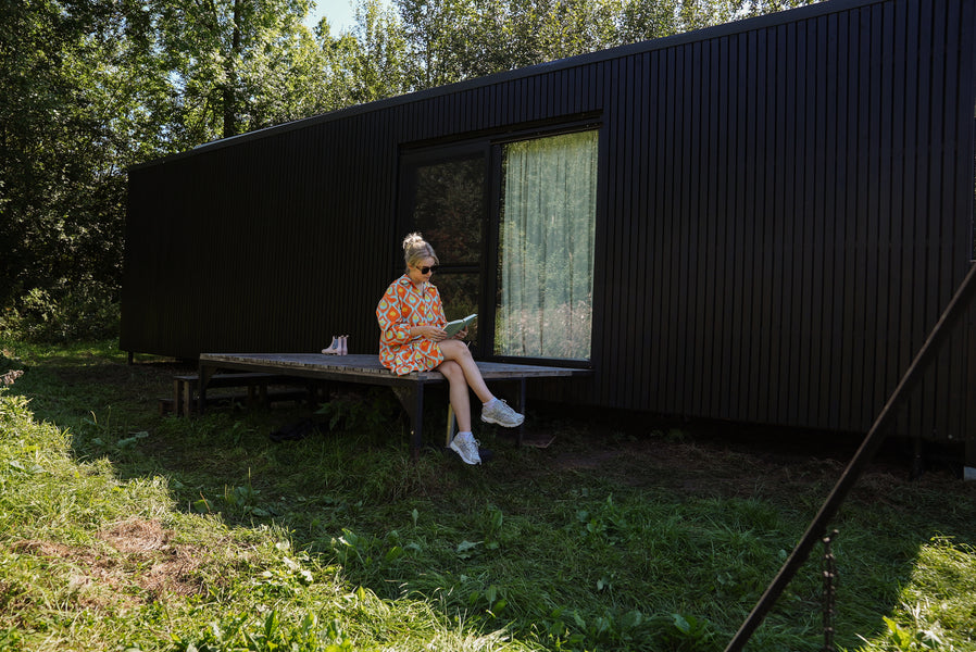 A woman is sitting on the edge of a wood deck reading a book with the black slow cabin finished with Rubio Monocoat DuroGrit is in the background.