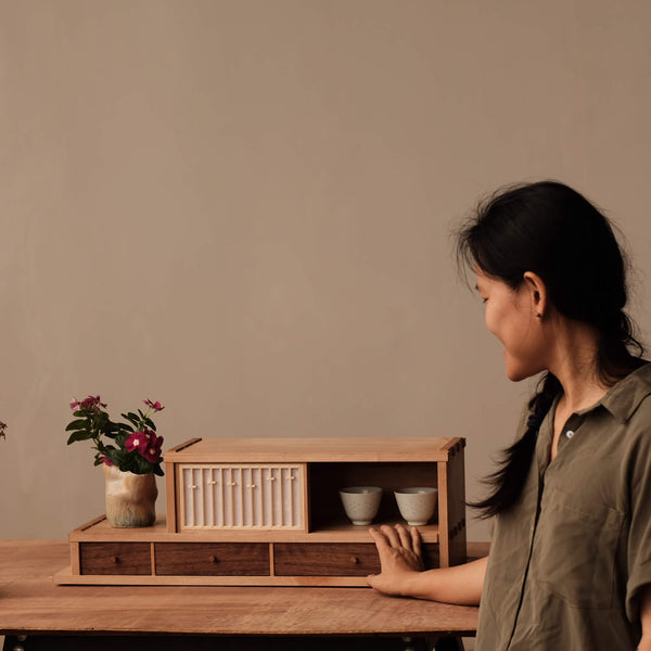 A woman is looking at a maple tea cabinet with two tea cups placed inside. Three walnut drawers are also on the bottom portion of the tea cabinet for storage.