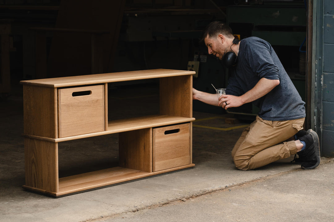 Woodworker applying finish to a handcrafted white oak bookcase with removable storage cubes, finished in Rubio Monocoat Oil Plus 2C Dunes, in a wood shop workshop.