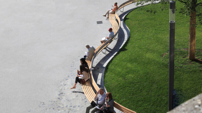 A wood bench curves around a green lawn and has people sitting on it.