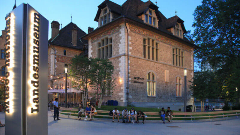 Curved benches with people sitting on them in front of the Swiss National Museum.