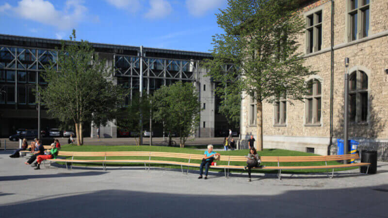 A wood bench with people sitting on it winds through a courtyard.