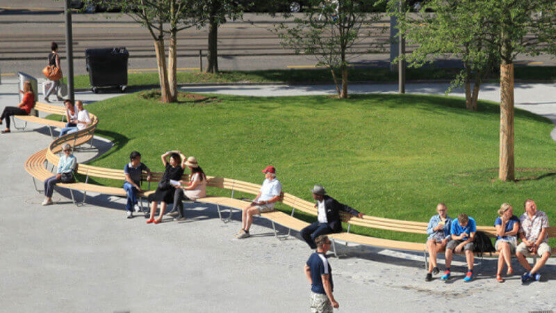 A curved wood bench with people sitting on it in front of a grass yard.