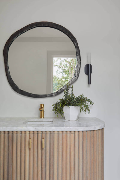 White oak bathroom vanity with a plant sitting on the marble counter top and round mirror above the faucet.