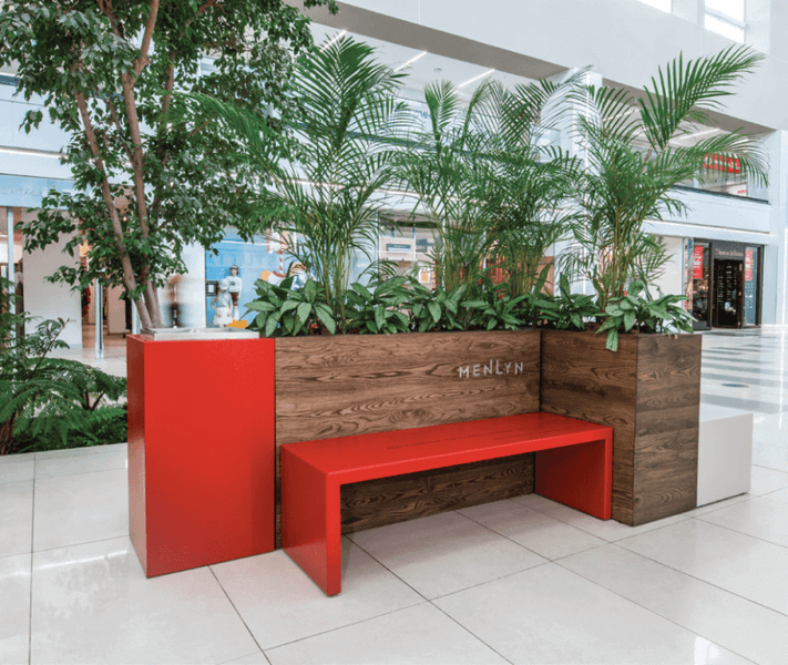 A brown wood planter box inside a mall with a bright red bench and trash can beside it.